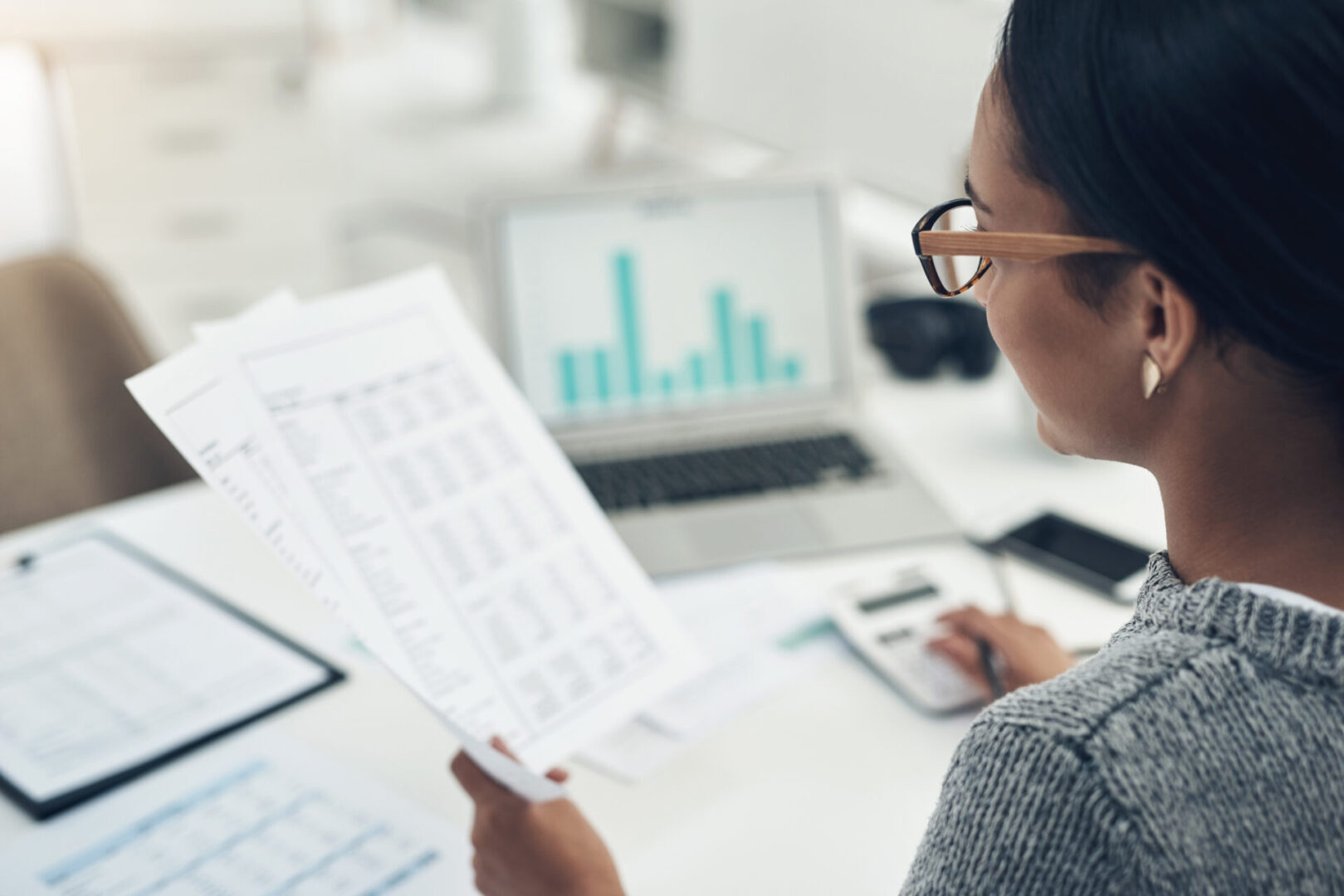Closeup shot of an unrecognisable businesswoman calculating finances in an office Person with glasses reviews documents and uses a calculator at a desk with a laptop showing a bar graph.
