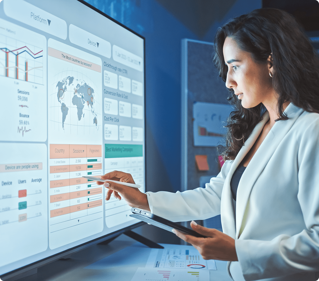 A woman in business attire interacts with a touchscreen displaying graphs and data analytics. She holds a tablet in her other hand.