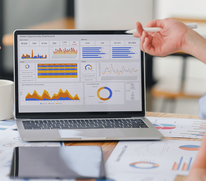 Laptop displaying a sales dashboard with various graphs and charts, surrounded by additional documents on a desk. A person is gesturing with a pen near the screen.