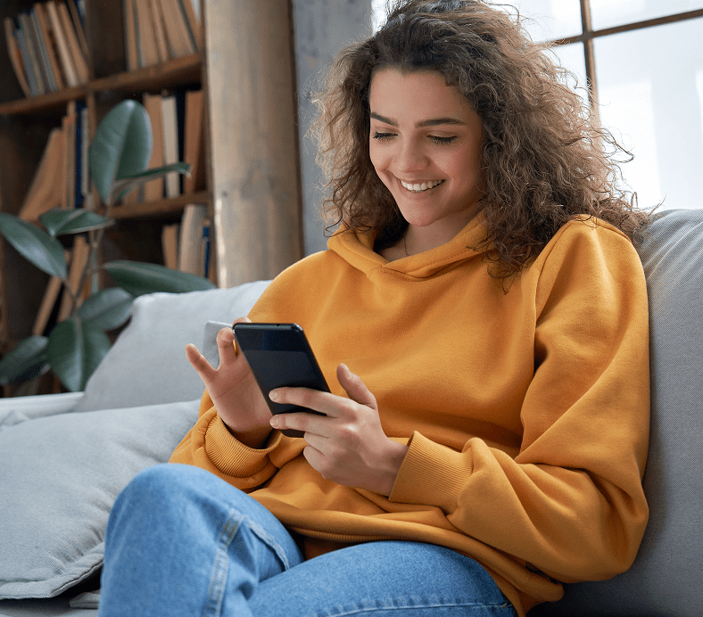 Woman in orange hoodie sits on a couch, smiling at her smartphone. There's a bookshelf and potted plant in the background.
