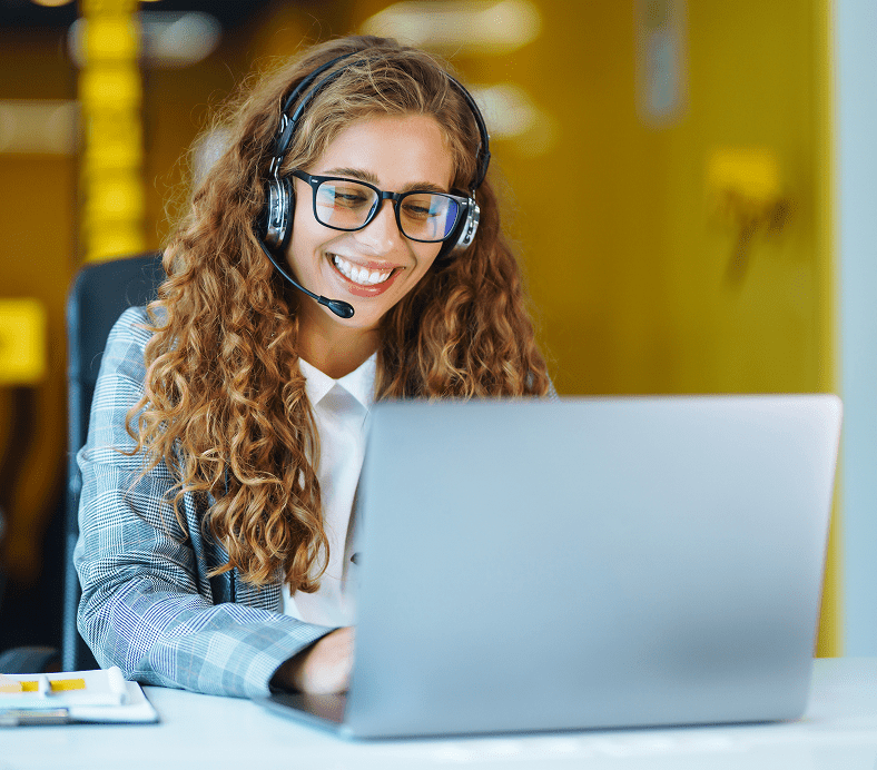 A person with curly hair and glasses is smiling while wearing a headset and using a laptop in an office setting.