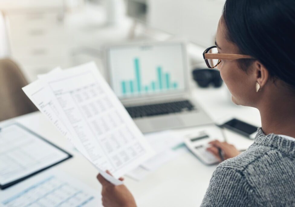Person with glasses reviews documents and uses a calculator at a desk with a laptop showing a bar graph.
