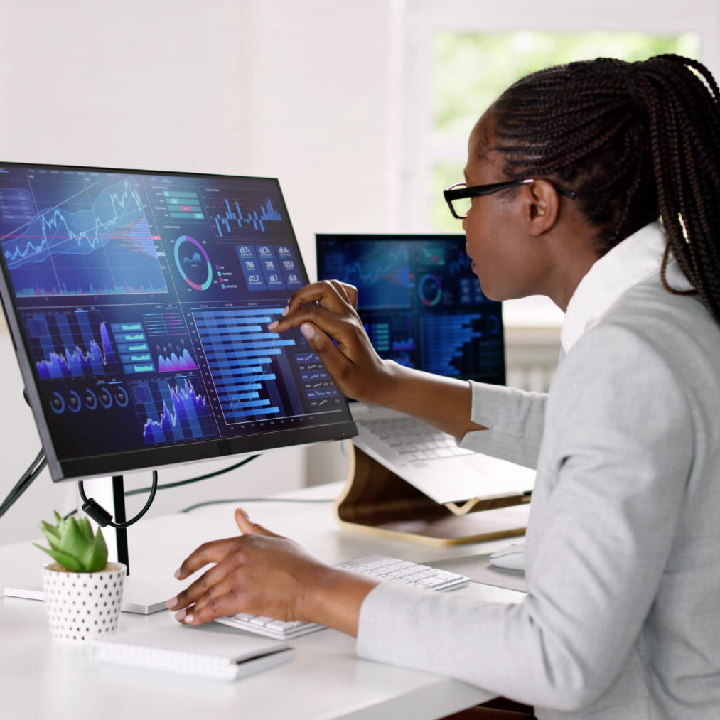 African Advisor Using KPI Dashboard A person in a gray blazer sits at a desk, analyzing data on a large computer monitor displaying graphs and charts.